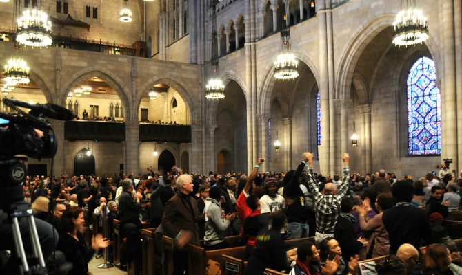 Bob-Avakia Cornel-West Dialogue Riverside-Church-Interior Crowd-in-pews-and-raised-fists-and-arms 11152014 Photo-Credit RevCom 670x400